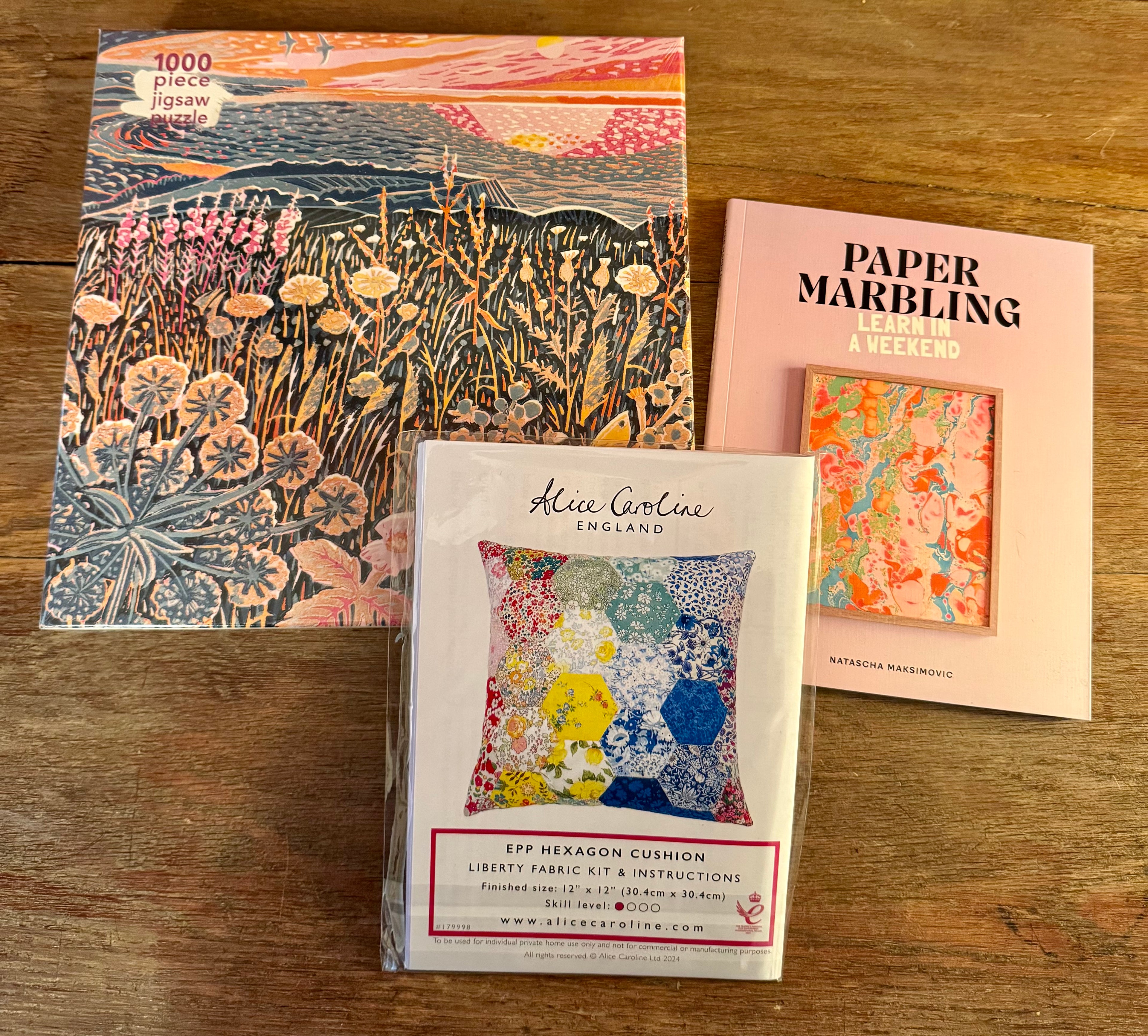 Three craft items on a wooden surface: a puzzle box, a fabric kit, and a paper marbling book.